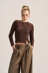 A model wearing a brown long-sleeve button-front crop top and high-waisted brown pleated trousers, standing against a light beige studio background.