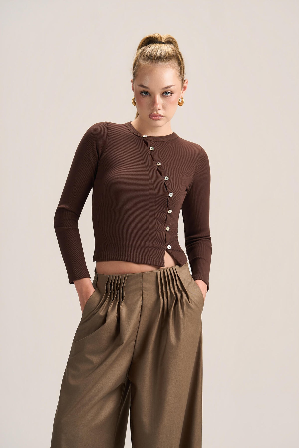 A model wearing a brown long-sleeve button-front crop top and high-waisted brown pleated trousers, standing against a light beige studio background.