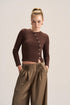 A model wearing a brown long-sleeve button-front crop top and high-waisted brown pleated trousers, standing against a light beige studio background.