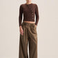 A model stands against a plain beige studio background wearing a dark brown ribbed long-sleeve wrap-style top with decorative buttons and high-waisted olive brown wide-leg trousers.