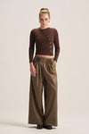 A model stands against a plain beige studio background wearing a dark brown ribbed long-sleeve wrap-style top with decorative buttons and high-waisted olive brown wide-leg trousers.