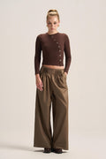 A model stands against a plain beige studio background wearing a dark brown ribbed long-sleeve wrap-style top with decorative buttons and high-waisted olive brown wide-leg trousers.