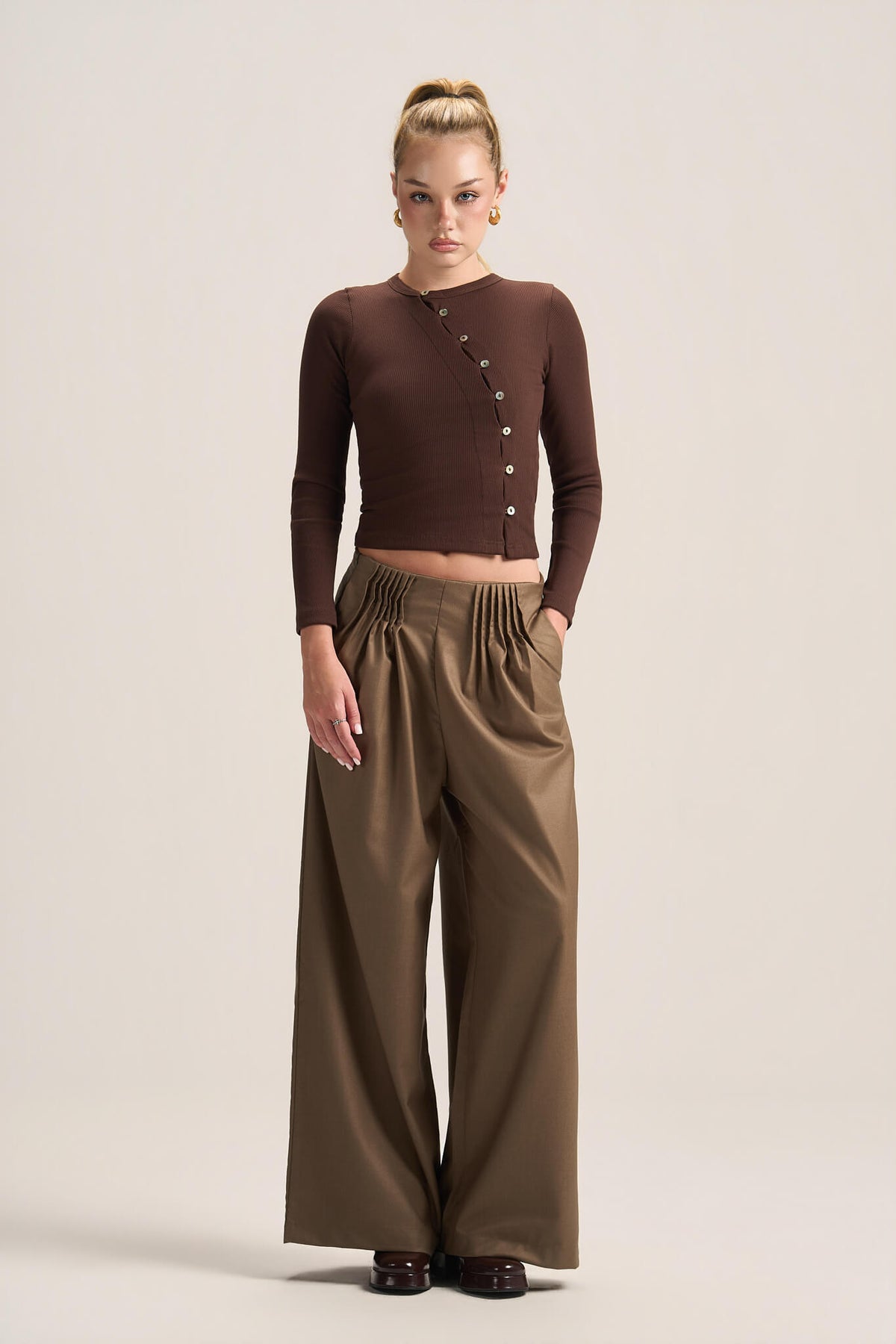 A model stands against a plain beige studio background wearing a dark brown ribbed long-sleeve wrap-style top with decorative buttons and high-waisted olive brown wide-leg trousers.