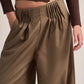 A close-up of a model wearing high-waisted, wide-leg trousers in a warm taupe-brown color with dramatic vertical pleats at the front waist, paired with a dark brown cropped long-sleeve top.