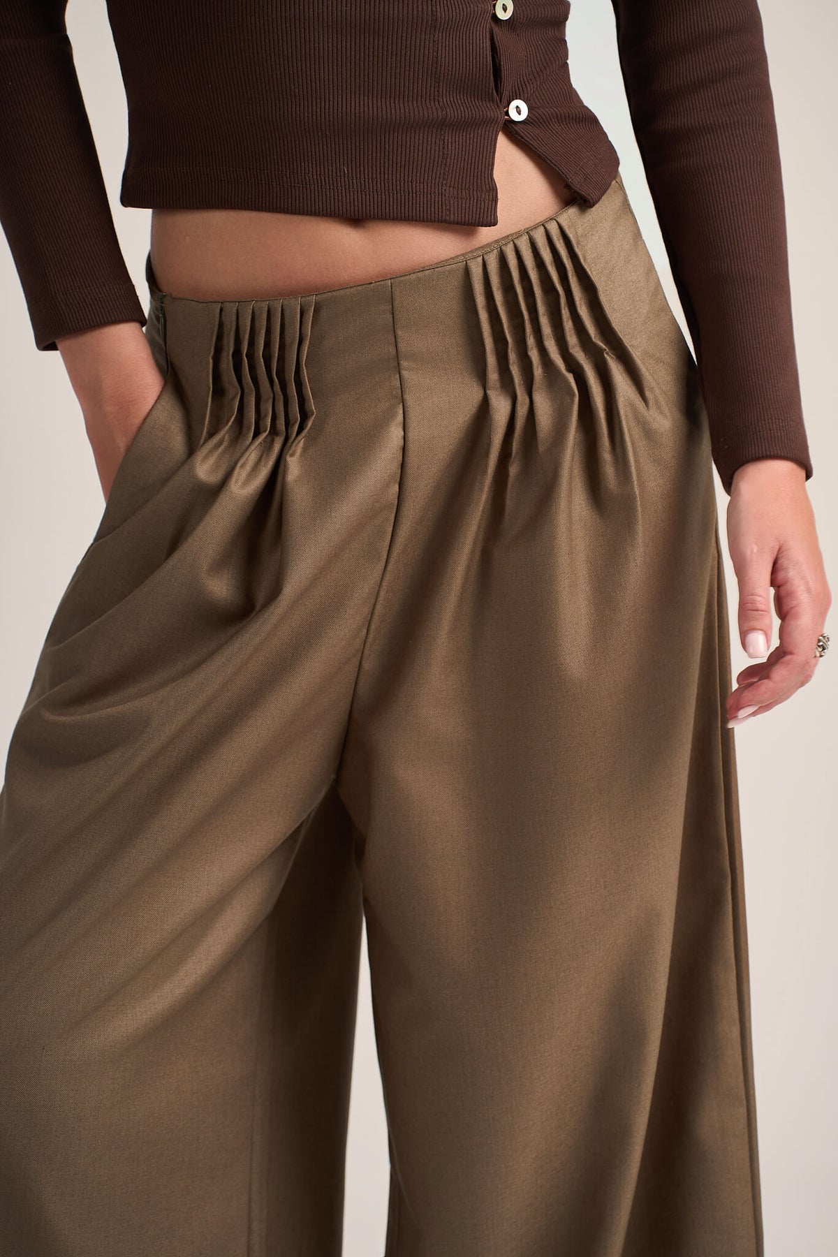 A close-up of a model wearing high-waisted, wide-leg trousers in a warm taupe-brown color with dramatic vertical pleats at the front waist, paired with a dark brown cropped long-sleeve top.