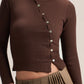A close-up of a model wearing a dark brown ribbed long-sleeve crop top with a buttoned asymmetrical neckline, paired with olive green pleated pants.