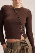 A close-up of a model wearing a dark brown ribbed long-sleeve crop top with a buttoned asymmetrical neckline, paired with olive green pleated pants.