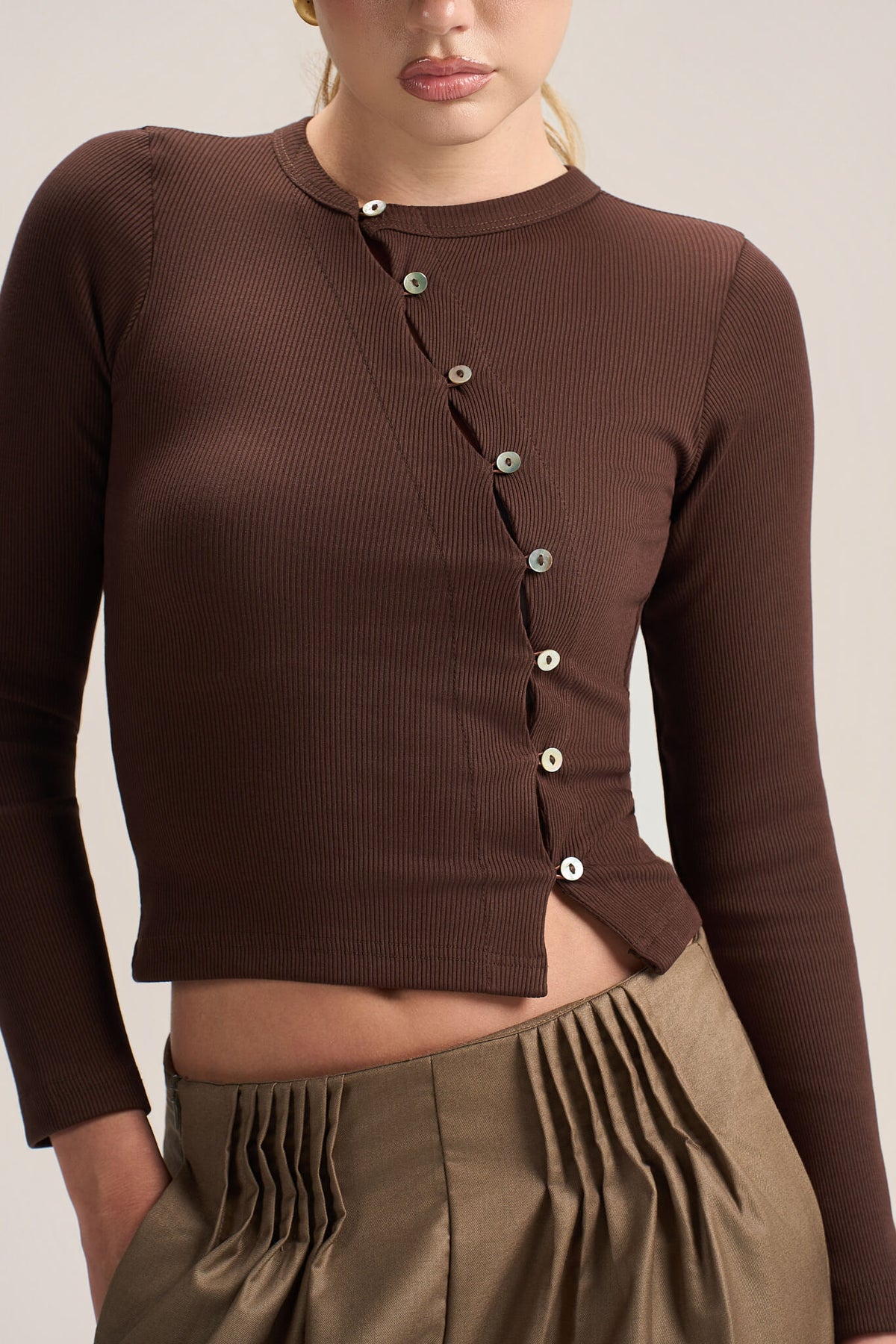 A close-up of a model wearing a dark brown ribbed long-sleeve crop top with a buttoned asymmetrical neckline, paired with olive green pleated pants.