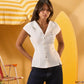 A woman modeling a white short-sleeve button-front top with a pointed collar, paired with dark jeans, posed in front of a yellow beach-themed backdrop with a striped umbrella and surfboard.