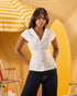 A woman modeling a white short-sleeve button-front top with a pointed collar, paired with dark jeans, posed in front of a yellow beach-themed backdrop with a striped umbrella and surfboard.