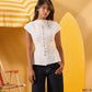 A woman modeling a white short-sleeve top with decorative metallic buttons down the front, paired with dark jeans, set against a warm yellow background with beach-themed props.