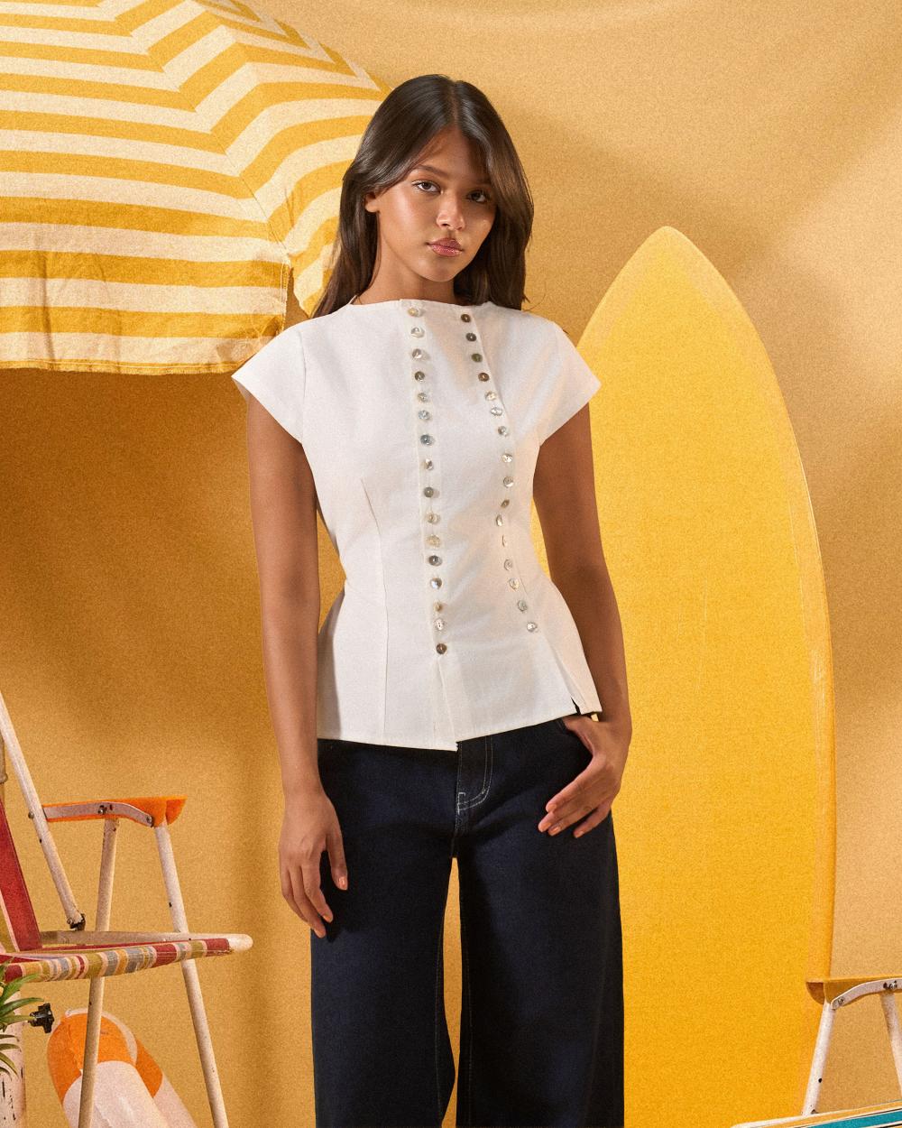 A woman modeling a white short-sleeve top with decorative metallic buttons down the front, paired with dark jeans, set against a warm yellow background with beach-themed props.