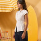 A woman modeling a white structured short-sleeve top with decorative buttons, paired with dark pants, set against a warm yellow beach-themed backdrop with a striped umbrella and surfboard.