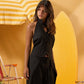 A woman modeling a black sleeveless halter-style top and matching black skirt against a warm yellow beach-themed backdrop with a striped umbrella and surfboard.