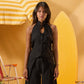 A woman stands in a studio setting wearing a black sleeveless halter-style top with a front tie and button detail, paired with matching black wide-leg bottoms. The background is warm yellow and orange with beach-themed props including a striped umbrella and surfboard.