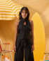 A woman stands in a studio setting wearing a black sleeveless halter-style top with a front tie and button detail, paired with matching black wide-leg bottoms. The background is warm yellow and orange with beach-themed props including a striped umbrella and surfboard.