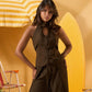 A woman models a sleeveless dark brown corset-style jumpsuit with a front keyhole tie and wide-leg pants, posed against a warm yellow beach-themed backdrop with a striped umbrella and surfboard.