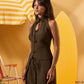 A woman modeling a sleeveless dark brown halter-neck dress with tie details, posed in front of a yellow surfboard and striped beach umbrella backdrop.