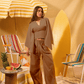 Woman in a brown outfit standing in a retro-style setting with a yellow and white striped umbrella, colorful chairs, and a pineapple.