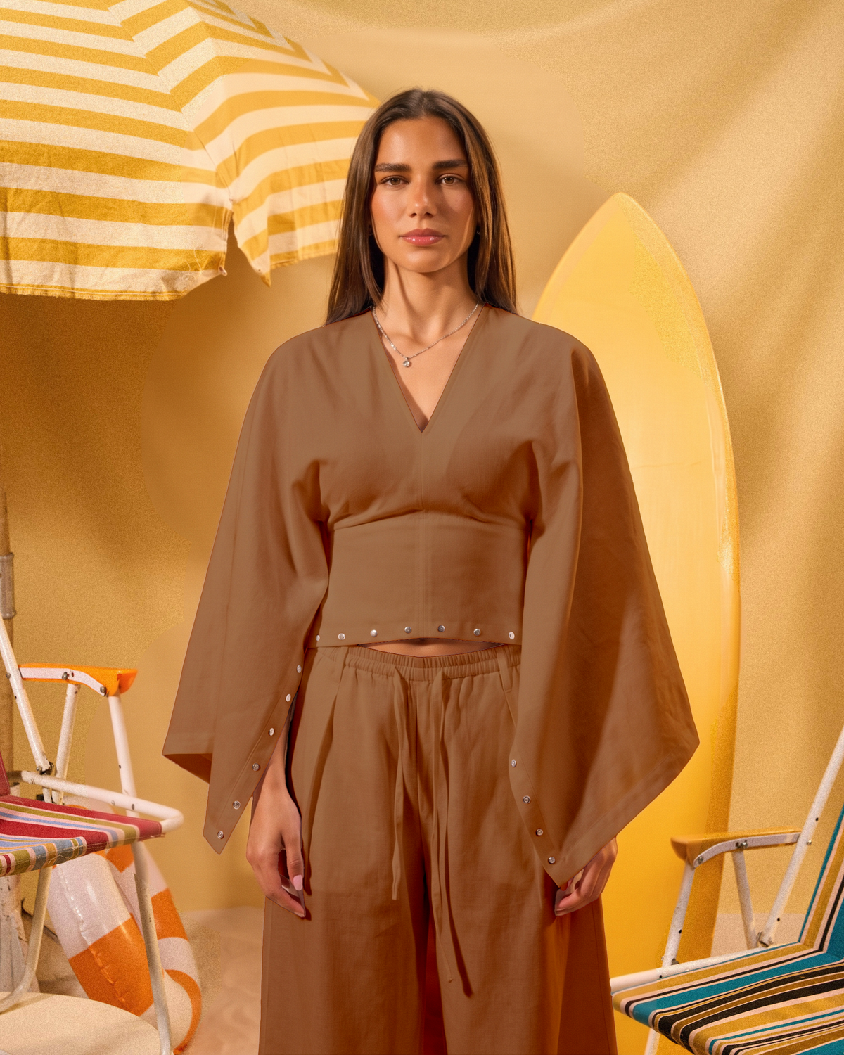 Woman in a brown outfit standing in front of a yellow umbrella and surfboard on a beach.