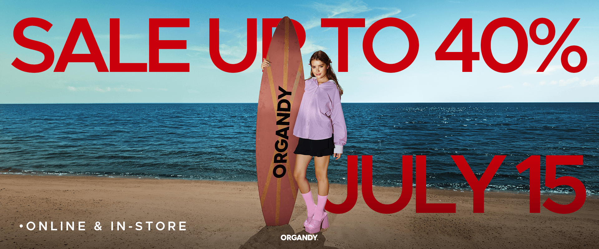 A promotional beach scene with a woman standing on sand holding a surfboard that reads ORGANDY. Large red text says SALE UP TO 40% and JULY 15, with ORGANDY branding at the bottom.slider_item_Jw8GkC
